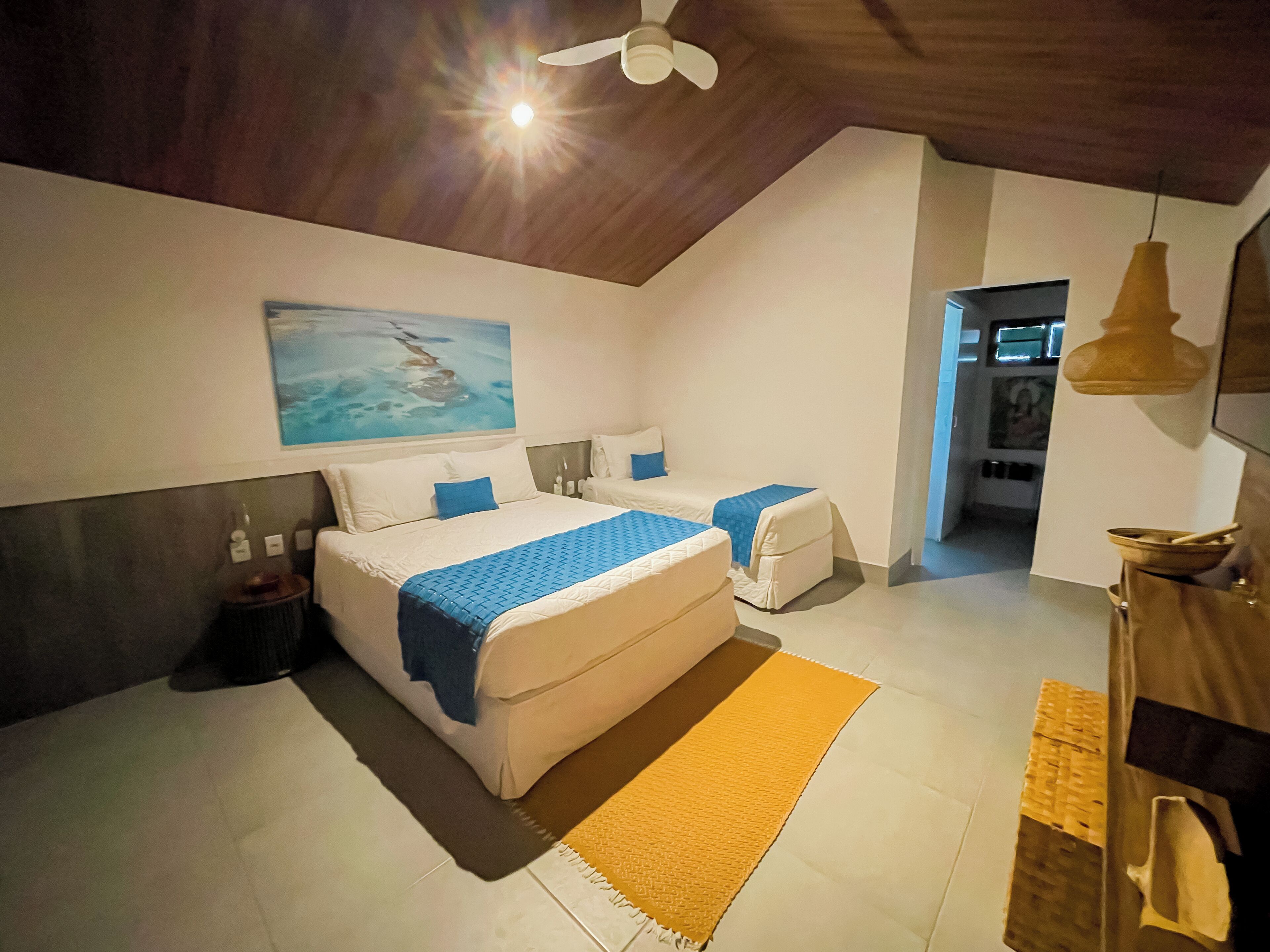 A bedroom with two beds, a ceiling fan, a painting, and a bathroom visible through an open door.