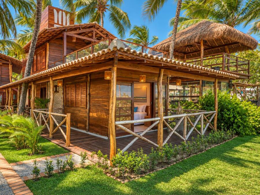 A wooden bungalow with a thatched roof, surrounded by lush greenery and palm trees.