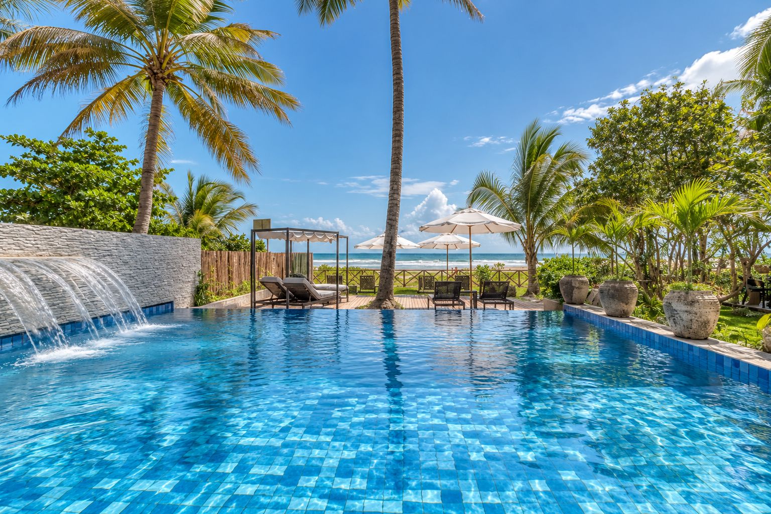 A clear blue pool with lounge chairs and umbrellas, surrounded by palm trees and a view of the ocean.