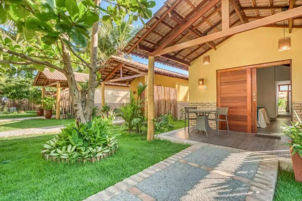 A covered outdoor area with a table and chairs, surrounded by greenery and a building with a wooden door.