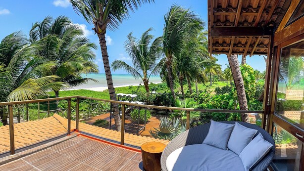 Un balcón con un sofá de mimbre, una mesa de madera y vistas a la playa y palmeras.