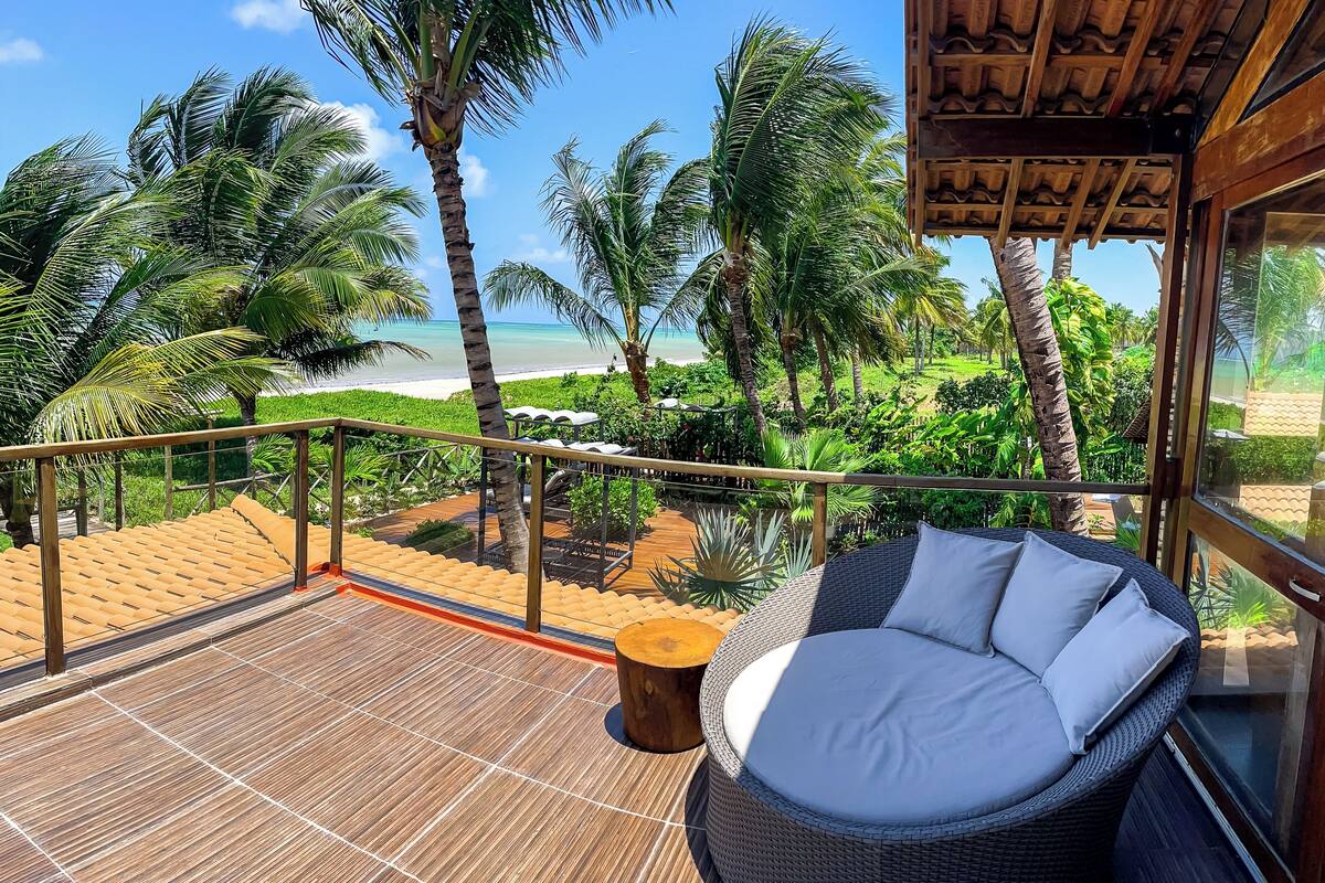 A balcony with a wicker sofa, a wooden table, and a view of the beach and palm trees.