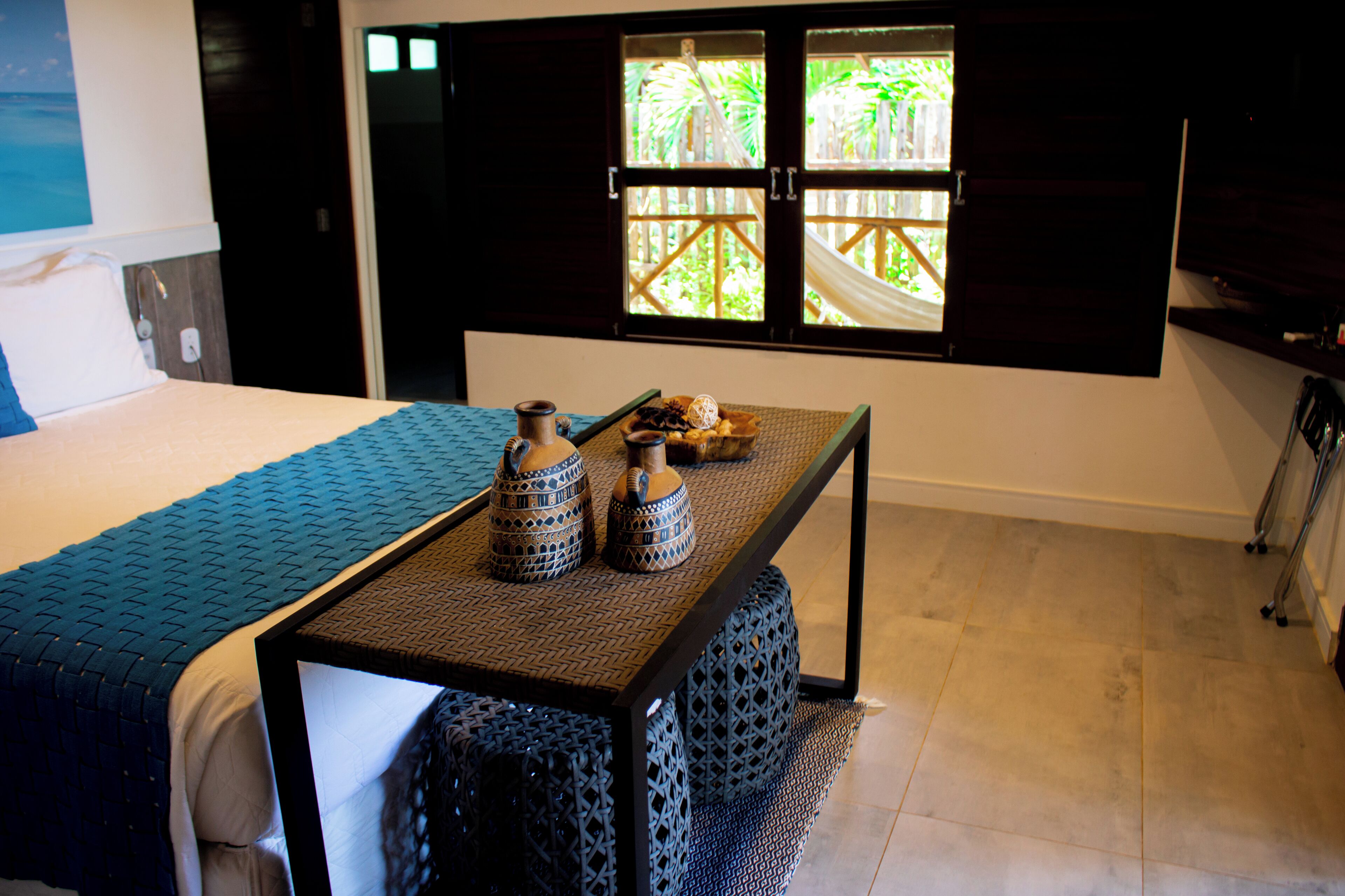 A hotel room with a bed, a table with decorative items, and a window with a view of greenery.