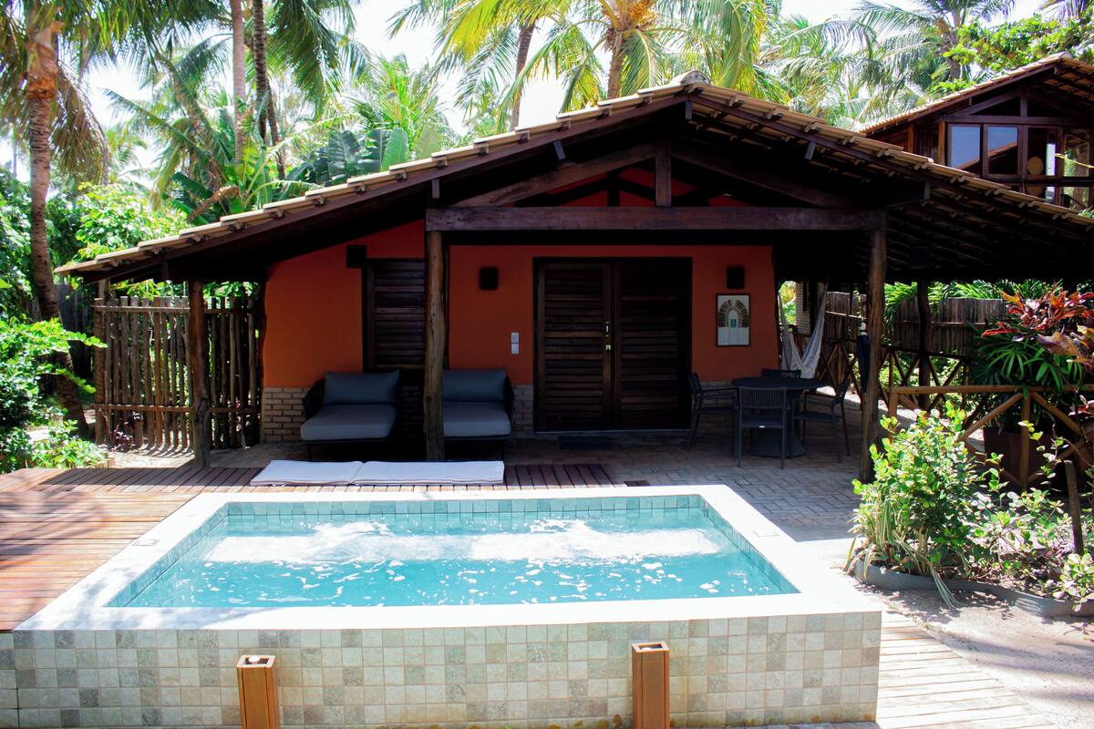 A pool with a wooden deck, a covered seating area, and a building with a sloped roof.