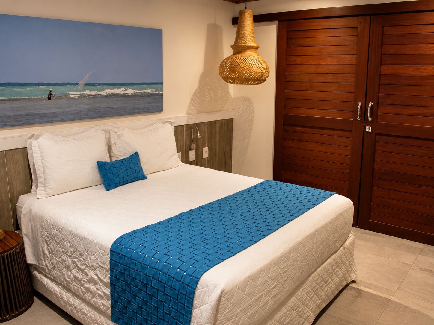 A bedroom with a bed, a blue quilt, a wooden wardrobe, and a beach view painting.