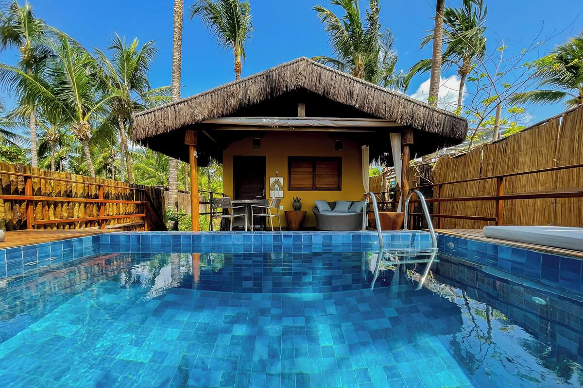 A pool area with a thatched-roof structure, a seating area, and a wooden fence.