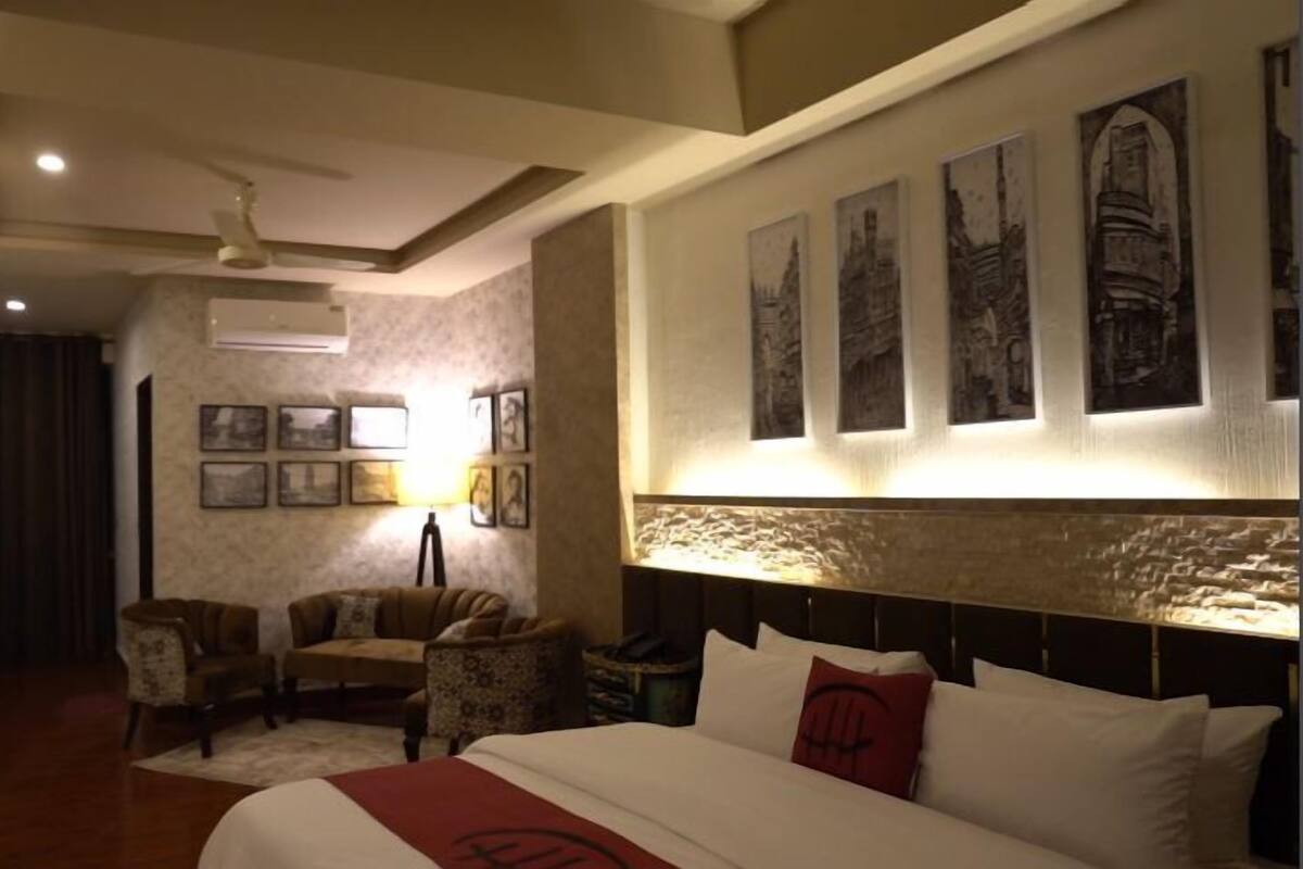A hotel room with a bed, two armchairs, a small table with a lamp, and a wall with framed pictures.