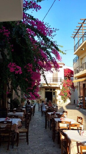 An outdoor café with tables and chairs, a cobblestone path, and buildings with balconies.