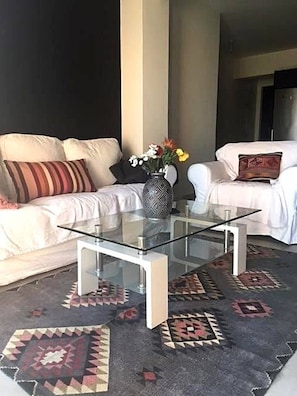 Una sala de estar con una mesa de centro de vidrio, dos sofás blancos, un tapete estampado y un jarrón con flores.