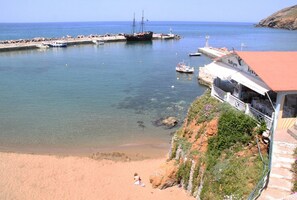 A coastal scene with a sandy beach, a rocky cliff with vegetation, a pier with boats, and a building with a red roof.