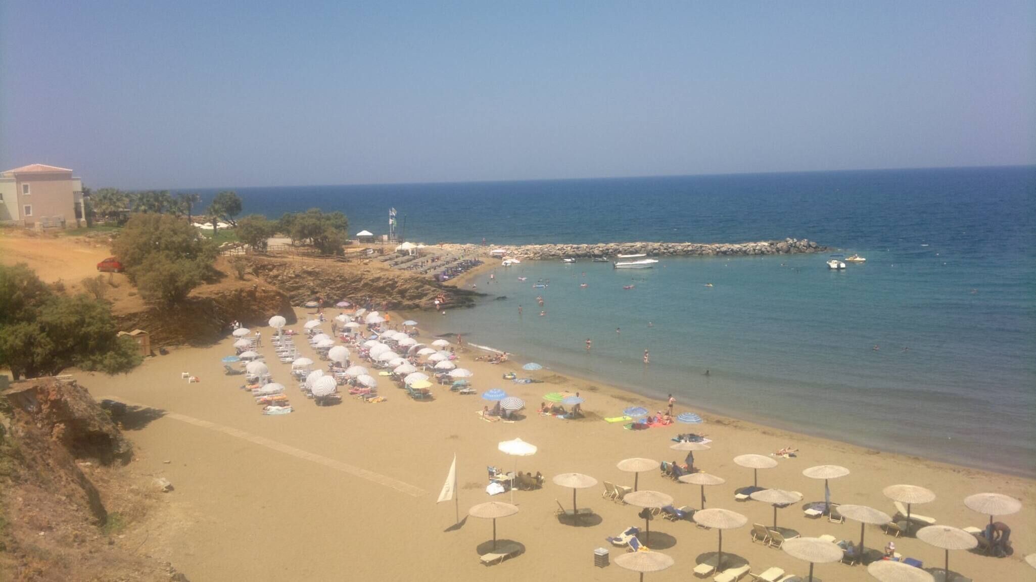 A beach with numerous umbrellas and sun loungers, a rocky shoreline, and a clear view of the sea.