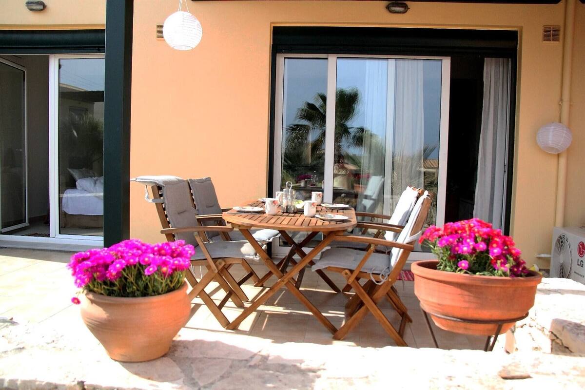 An outdoor seating area with a wooden table and chairs, two potted plants with pink flowers, and a sliding glass door leading to a covered patio.