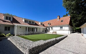 A courtyard with a green lawn, a stone wall, and a building with red-tiled roofs and white walls. - Chambre D'hôtes - Gary Player (AIX EN ISSART)