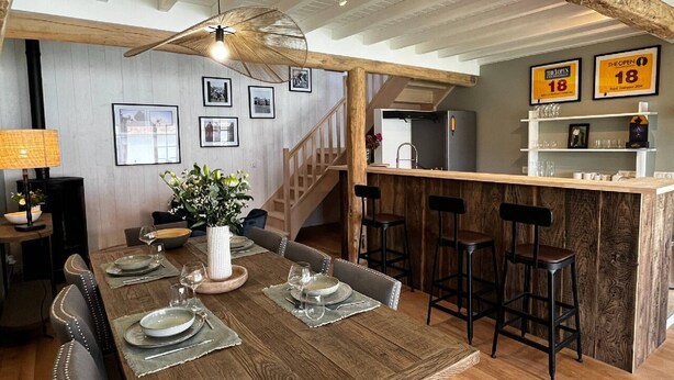 A dining area with a wooden table set for four, a bar area with stools, and a staircase leading to an upper level. - Chambre D'hôtes - Gary Player (AIX EN ISSART)