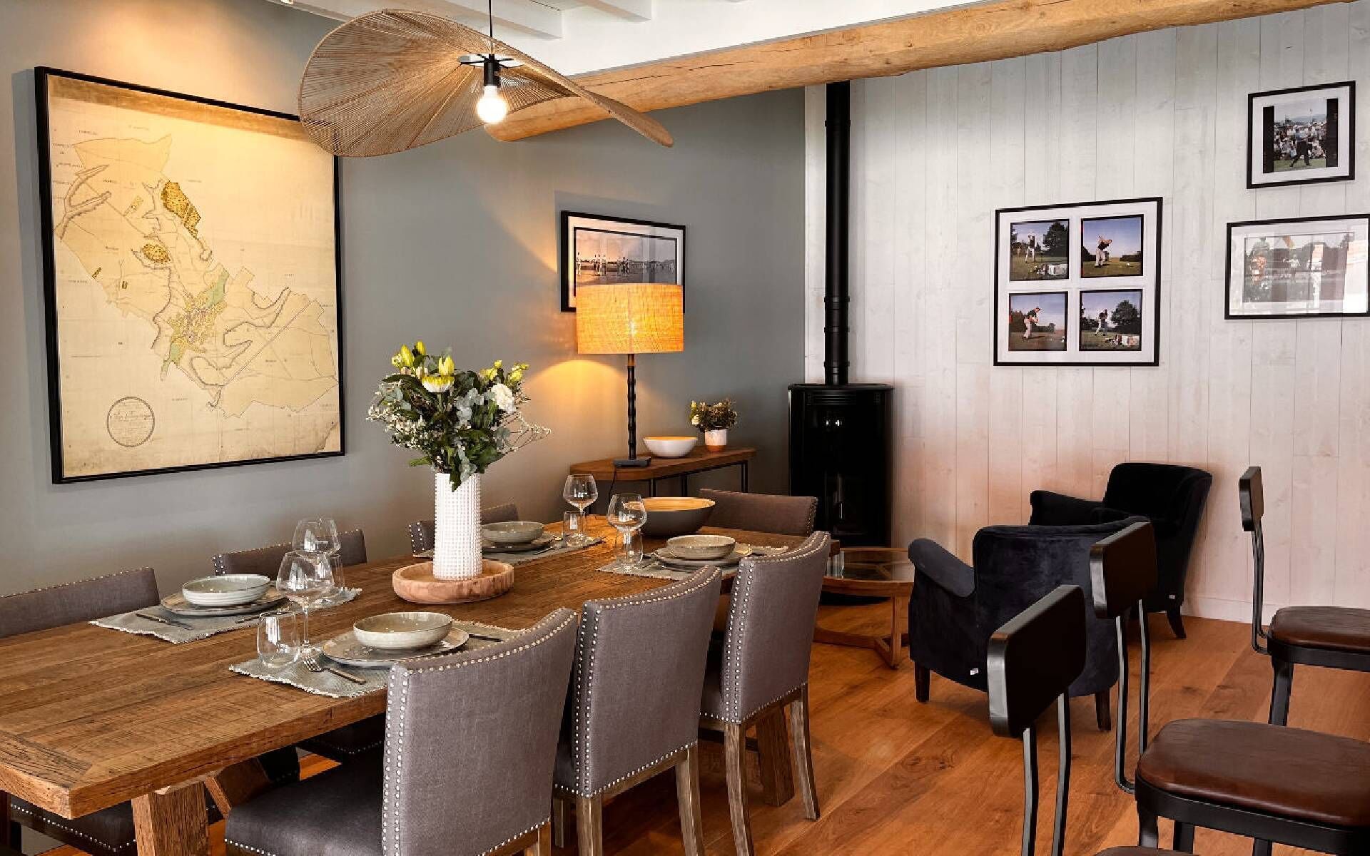 A dining area with a wooden table set for six, surrounded by upholstered chairs. A large map is displayed on the wall, and a lamp is positioned above the table.