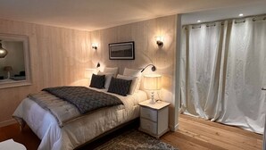 A bedroom with a bed, bedside lamps, a nightstand, a mirror, and a window with curtains. - Chambre D'hôtes - Gary Player (AIX EN ISSART)