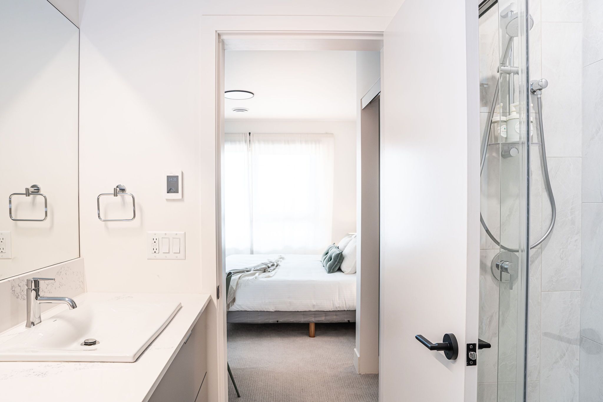 A modern bathroom with a white sink, a glass shower enclosure, and a bed with a white mattress and gray pillows in the adjacent room.