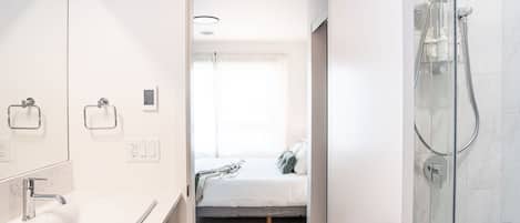 A modern bathroom with a white sink, a glass shower enclosure, and a bed with a white mattress and gray pillows in the adjacent room.