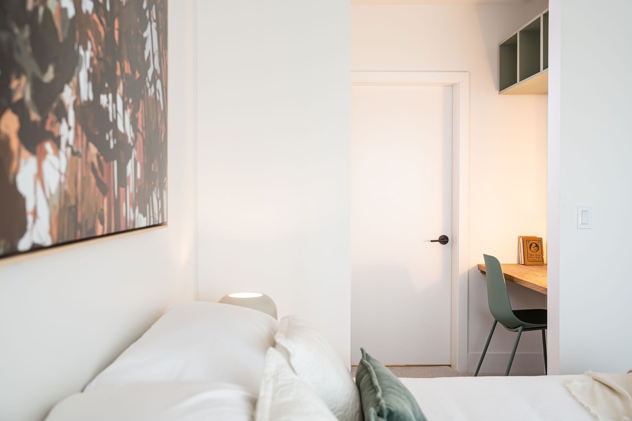 A bedroom with a bed, a desk with a chair, a door, and a wall-mounted shelf.