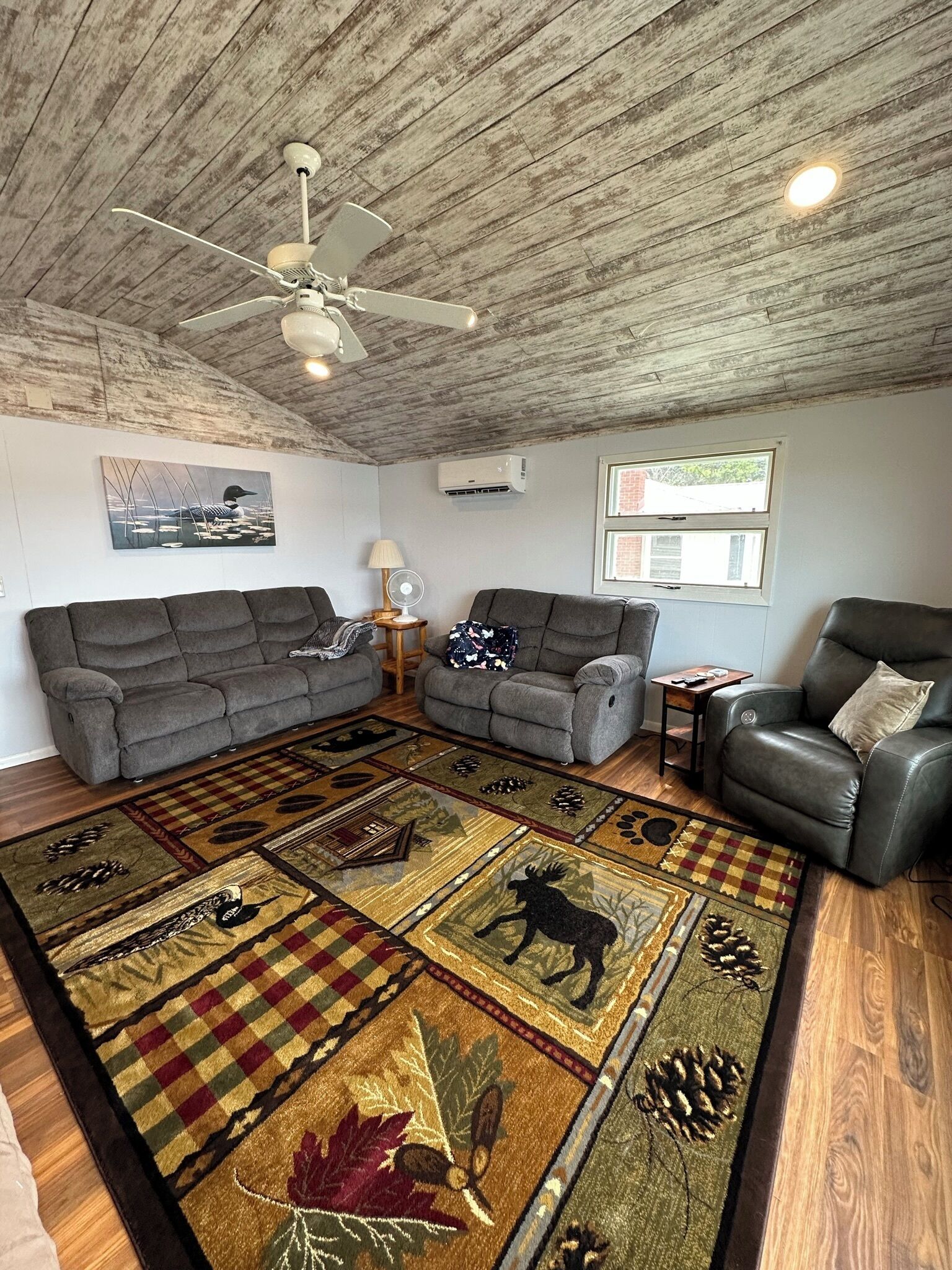 A living room with two grey sofas, a leather armchair, a patterned rug, a ceiling fan, and a window with a view.