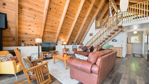 A spacious loft-style living area with a high ceiling, wooden beams, a leather sofa, a rocking chair, and a staircase leading to an upper level. - Moonlight Shores Lakefront (Fawnskin)