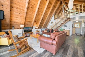 A spacious loft-style living area with a high ceiling, wooden beams, a leather sofa, a rocking chair, and a staircase leading to an upper level. - Moonlight Shores Lakefront (Fawnskin)