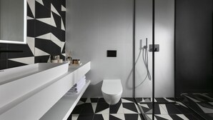Een moderne badkamer met een zwart-wit betegelde vloer in geometrisch patroon, een wit toilet, een glazen douchecabine en een minimalistische wastafel met spiegel en toiletartikelen.