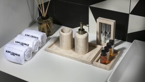 Une salle de bain avec un lavabo blanc, deux serviettes roulées étiquetées « King George Suites », un distributeur de savon, un récipient en verre contenant des bâtonnets de rotin et une petite étagère avec des flacons.