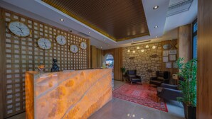 A reception area with a marble counter, wooden panel wall with clocks, stone wall with framed certificates, and leather seating. - Ornella Hotel (Edremit)