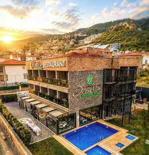 A hotel with a restaurant on the ground floor, a pool area, and a view of a hillside town. - Ornella Hotel (Edremit)