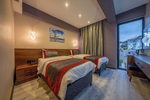 Habitación de hotel con dos camas, un escritorio, una silla, una mesita de noche y un ventanal con vistas a edificios.