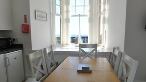 Un coin repas avec une table en bois et des chaises blanches, une cuisine équipée d’un four à micro-ondes et de grandes fenêtres offrant une vue dégagée.