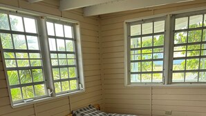 A room with two beds, each with a checkered bedspread, large windows with a view of greenery, and a tiled floor.
