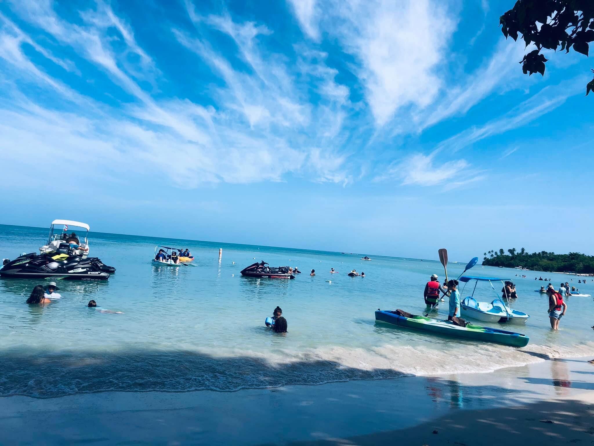 Ein Strand mit klarem, blauem Wasser, Booten und Menschen, die sich im Wasser vergnügen.
