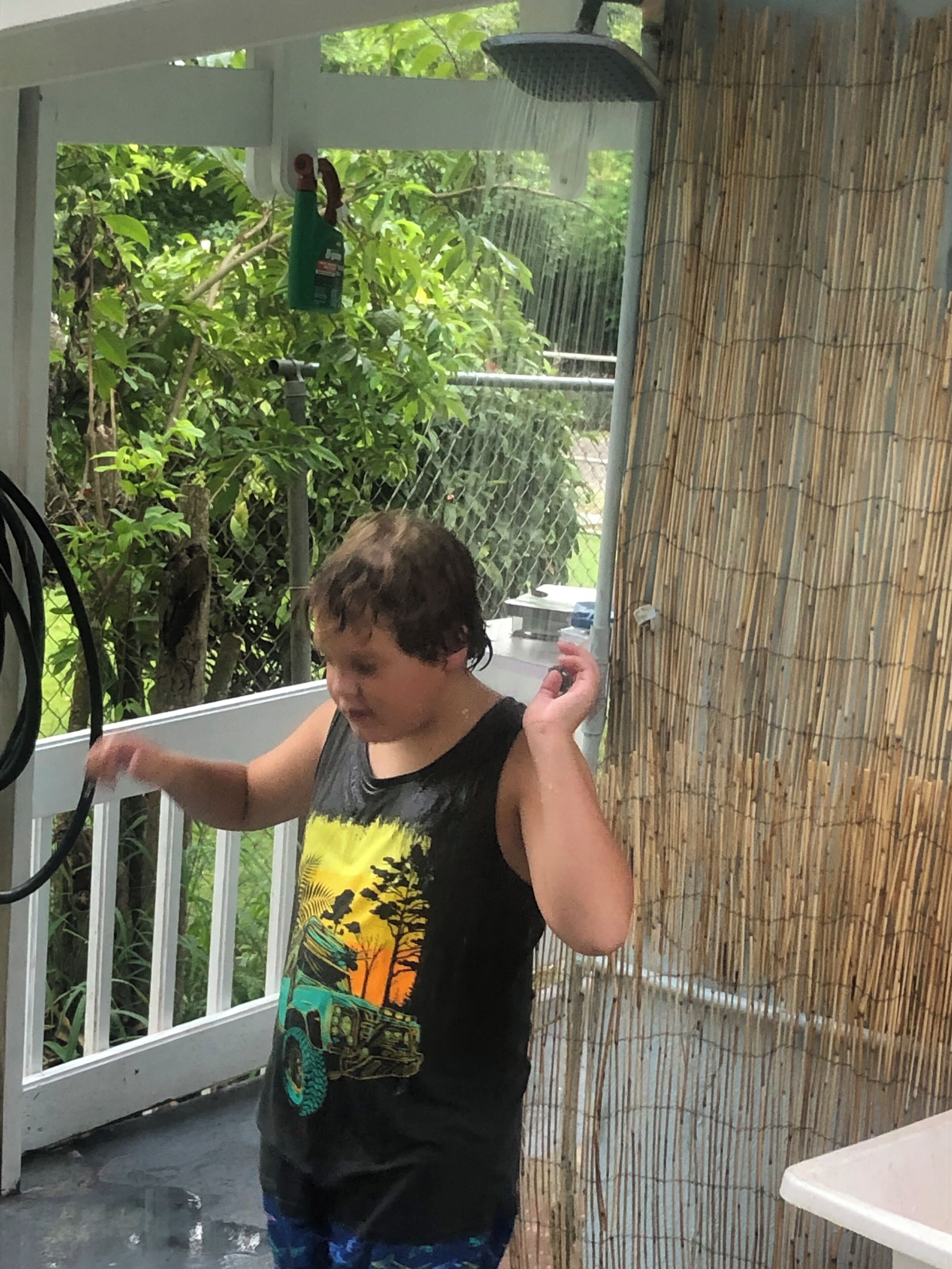 A child standing on a porch holding a hose, with a bamboo fence and greenery in the background.