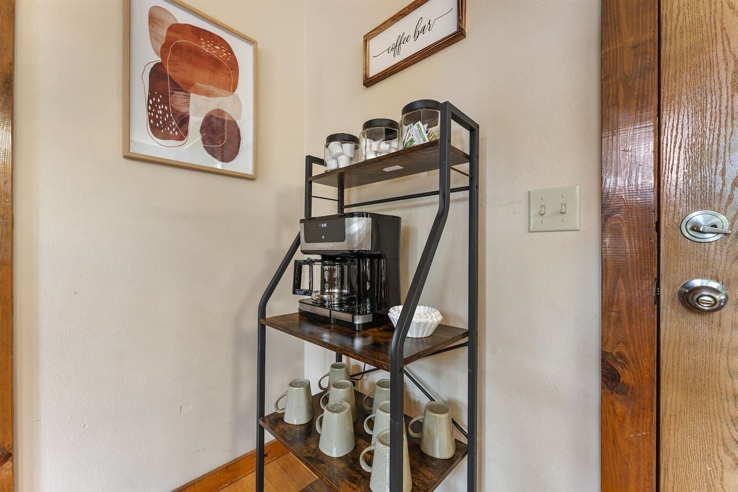 A coffee machine on a shelf with mugs below it, a coffee bar sign, and a framed picture on the wall.