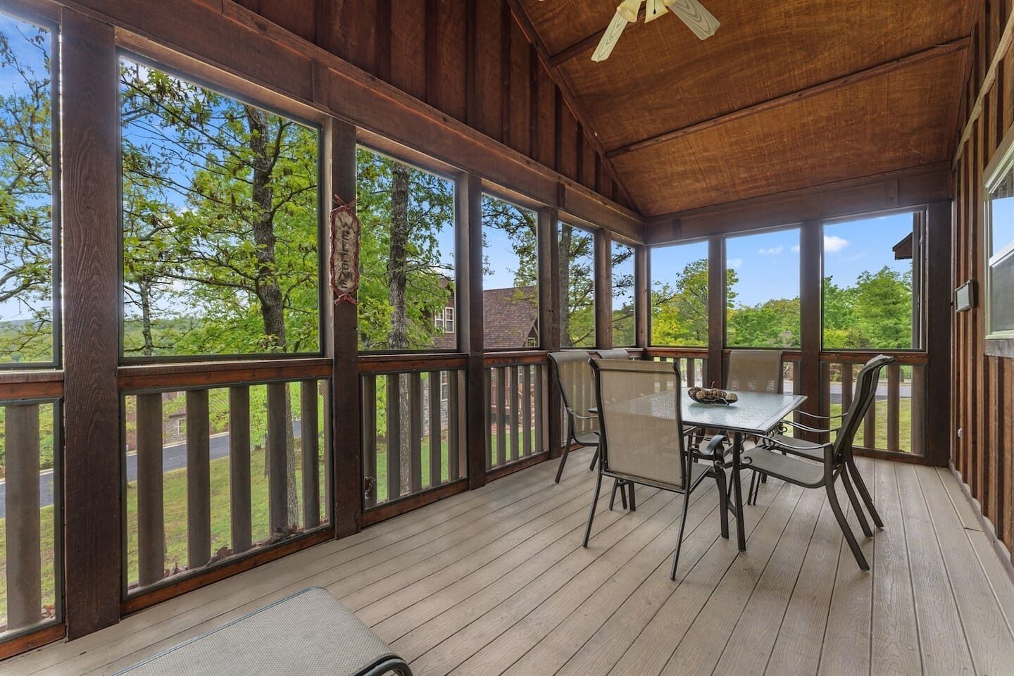 A covered outdoor deck with a table and chairs, large windows, and a wooden ceiling.