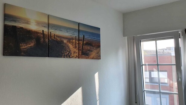 A room with a large wall-mounted artwork depicting a beach scene, a window with blinds, and a door. - Room-cosy Room In Antwerpen (Mortsel)