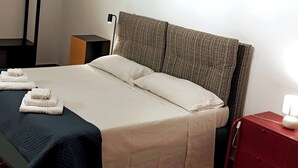 A neatly made bed with white linens and a dark blue bedspread, a red nightstand with a lamp, and a grey headboard. - Holiday Home Sofia Palermo (Palermo)