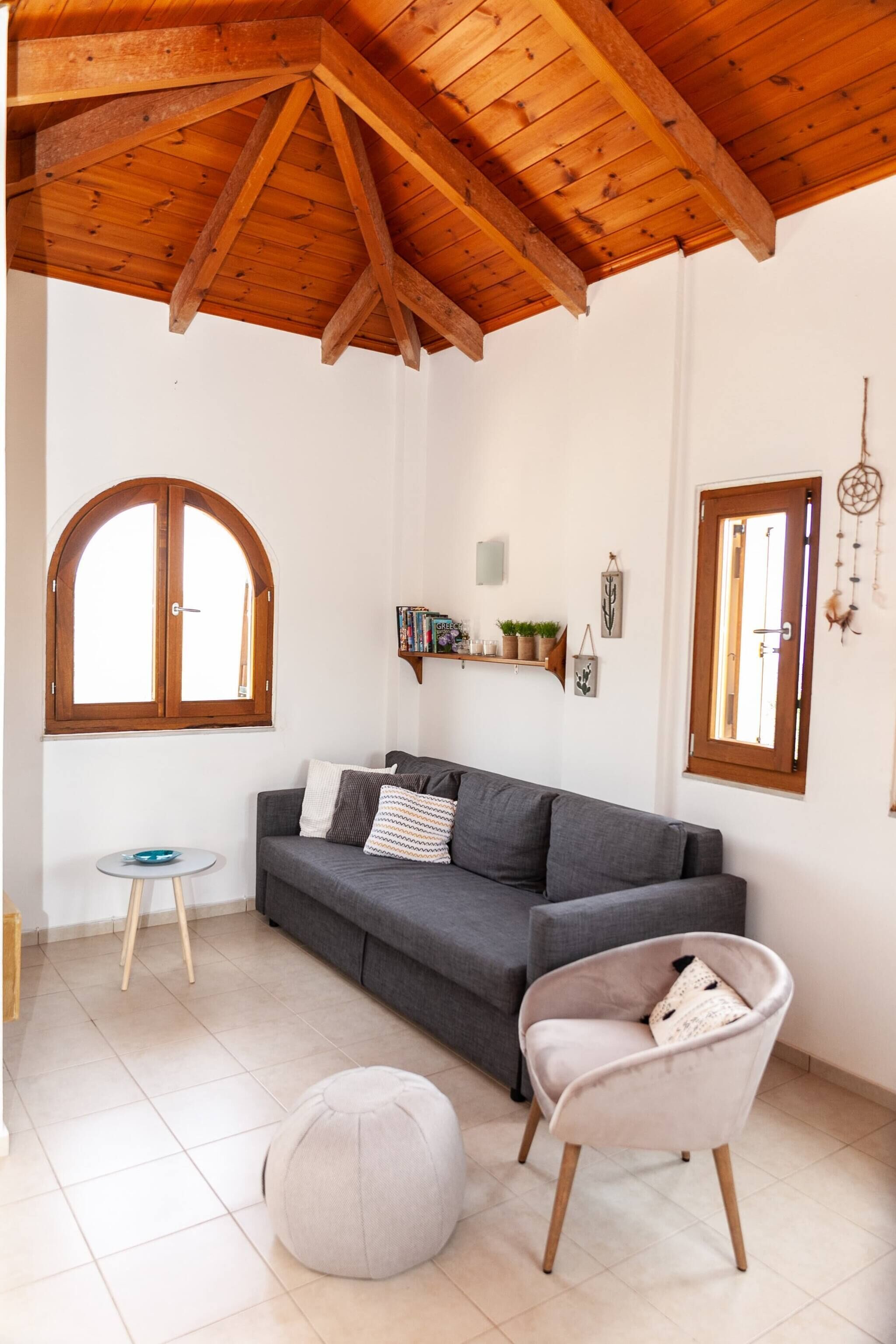 A cozy room with a wooden ceiling, a grey sofa, a small round ottoman, and a chair with a cushion.