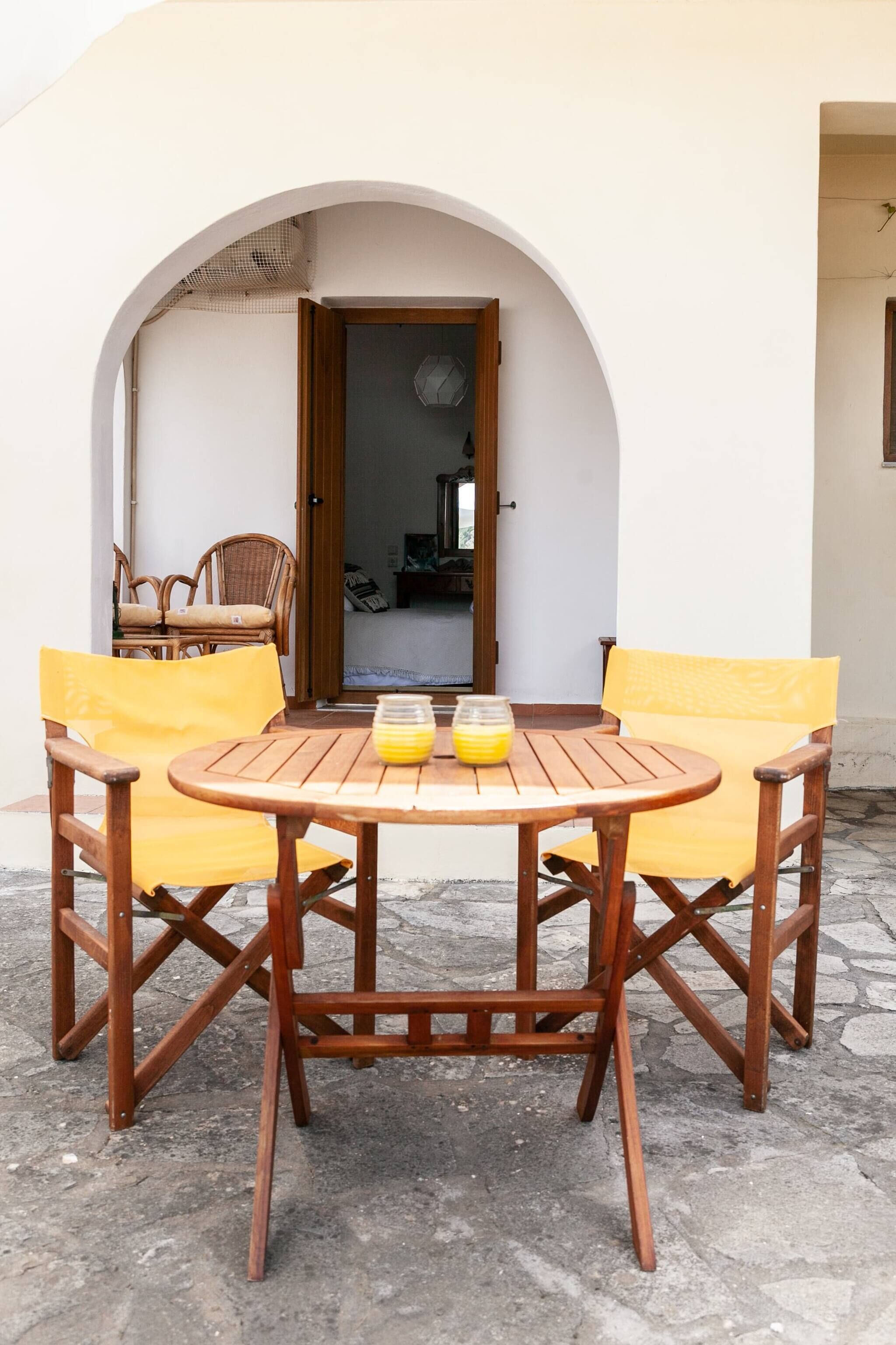 A wooden table with yellow cushions, a wicker chair, and a doorway leading to a room.
