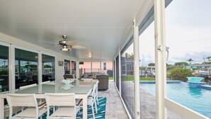 Un patio couvert avec une table et des chaises en verre, un ventilateur de plafond et une vue sur une piscine.