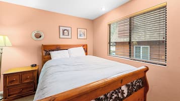 A bedroom with a wooden bed, a nightstand, a lamp, and a window with blinds.