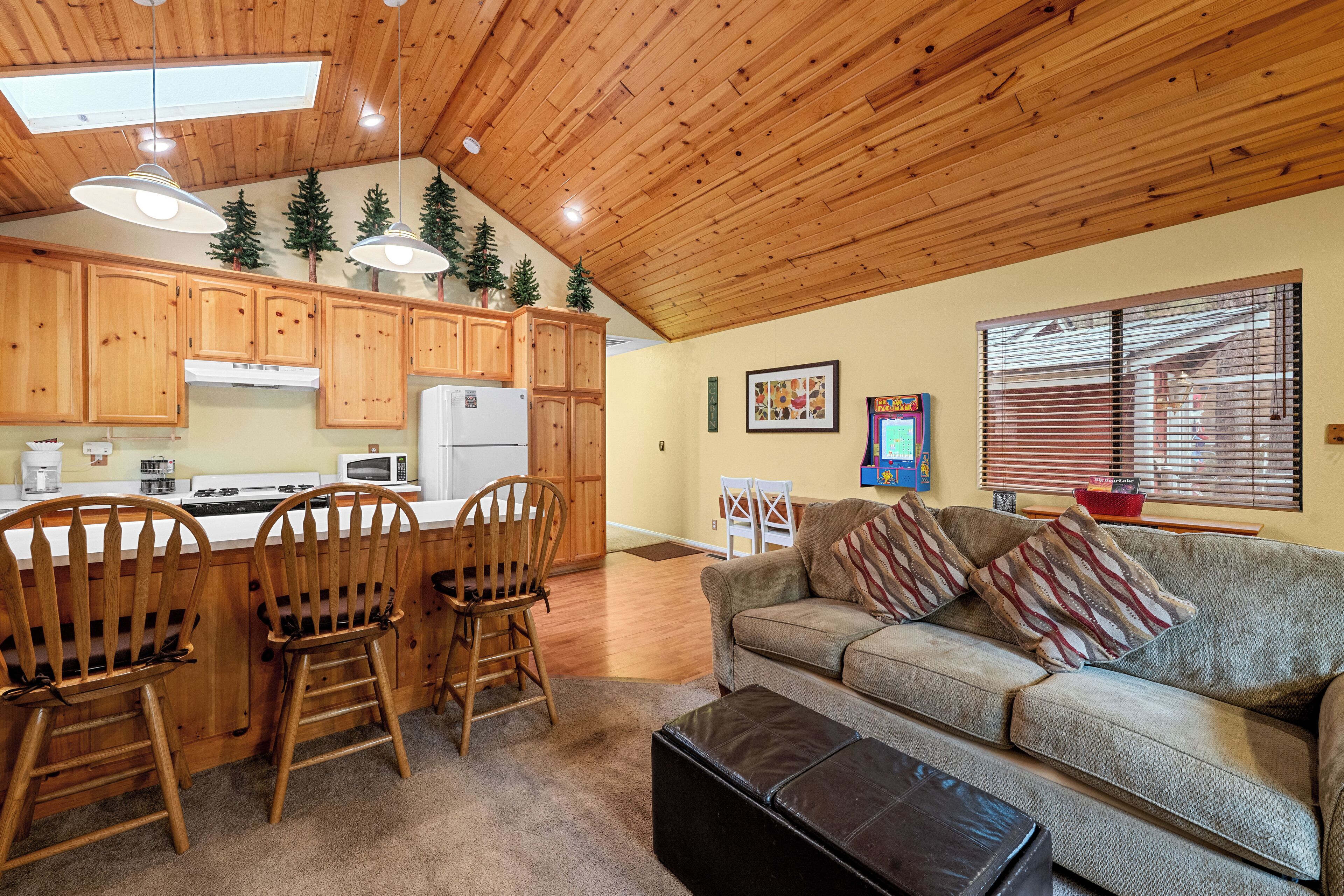 A cozy living room with a sofa and chairs, wooden cabinets, a kitchen area with a refrigerator, and a window with blinds.