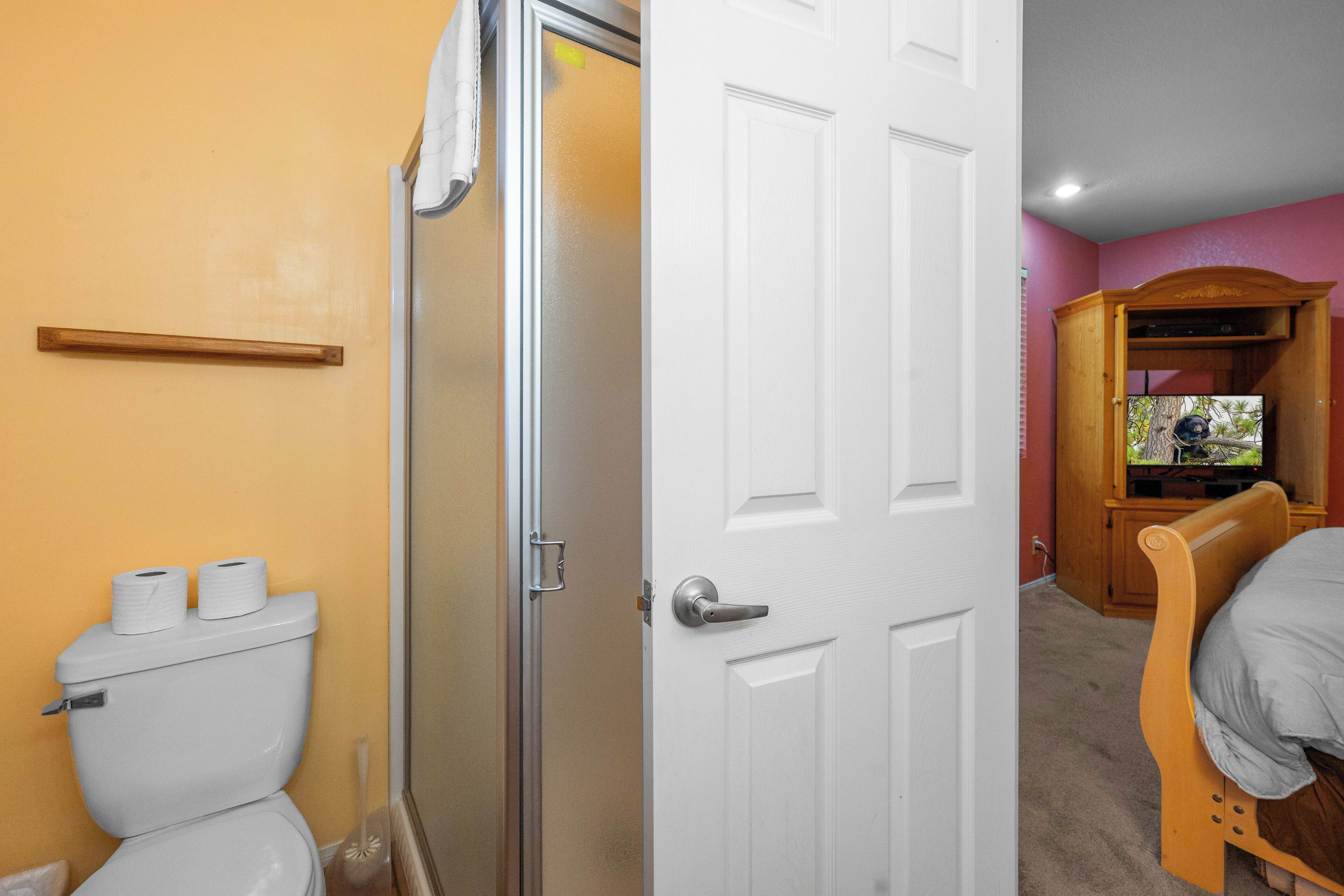 Un baño con inodoro, ducha y puerta que da a un dormitorio con cama y televisor.