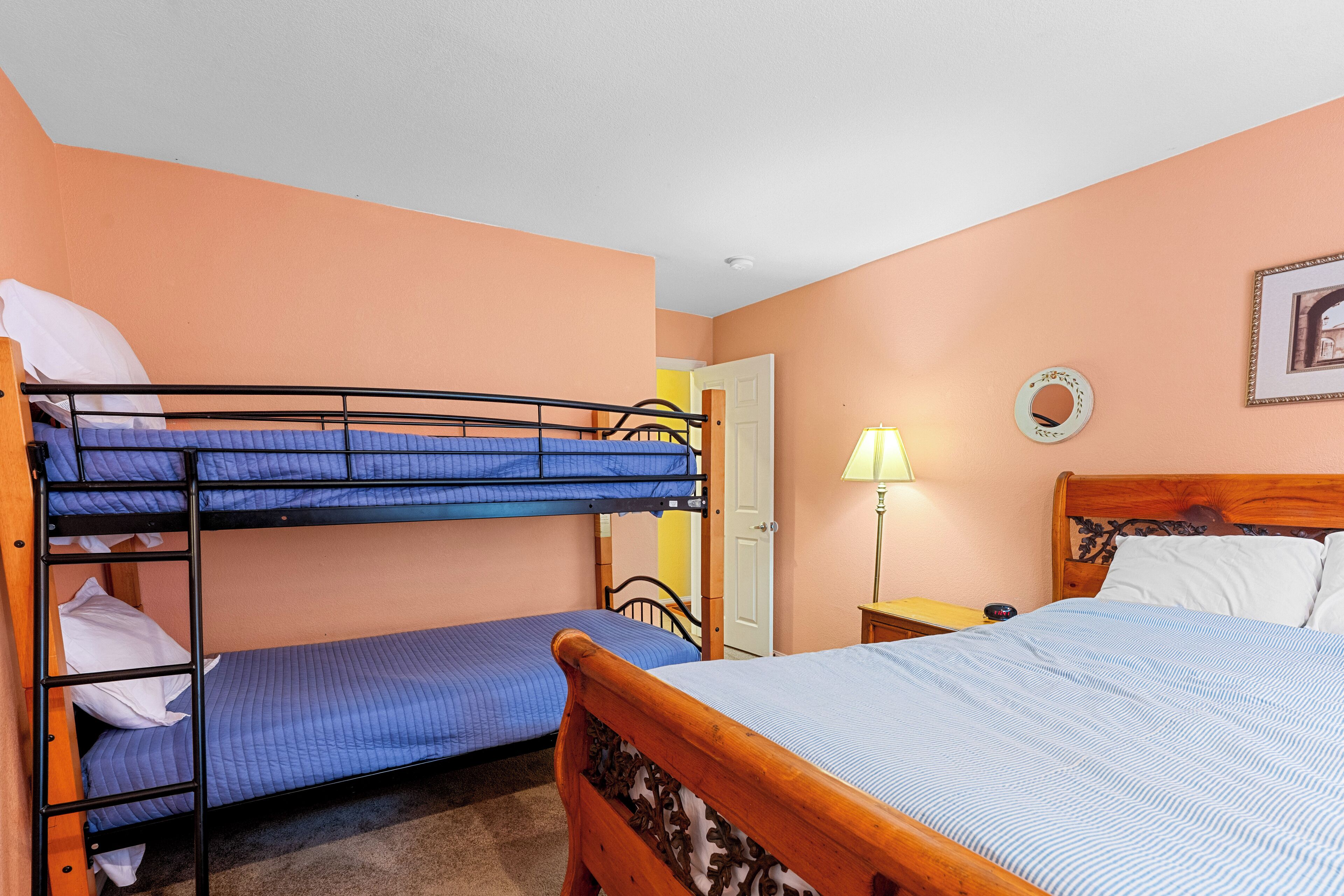 Quarto com beliches, uma cama de madeira, uma mesa de cabeceira, um abajur e um quadro na parede.