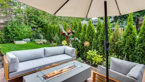 A modern outdoor seating area with a gray sofa and a glass-top table with a built-in fire feature, under a large umbrella. - Newly Built Maple Valley Vacation Rental w/ Patio! (Maple Valley)
