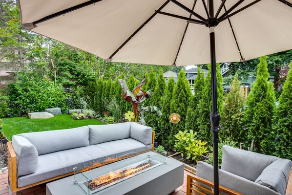 A modern outdoor seating area with a glass-top table, a grey sofa, and a wooden bench under a large umbrella.