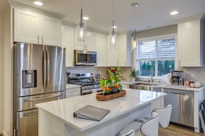 A modern kitchen with stainless steel appliances, a central island with seating, pendant lighting, and a view of the outdoors through a window.