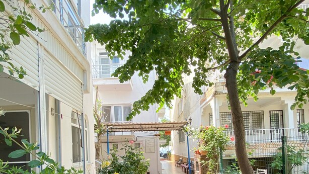 A courtyard with a tiled walkway, surrounded by buildings, greenery, and a tree providing shade. - Sedef Pansiyon Apart Otel Sarkoy (Sarkoy)